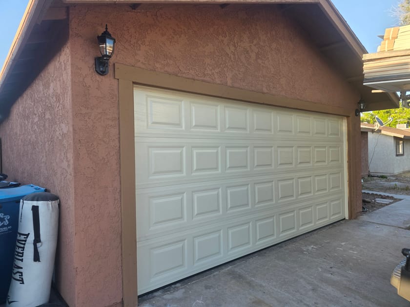 A cream double garage door beside a wall lamp on a tan stucco home.