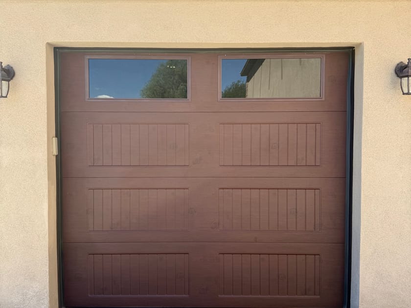 A dark wood look garage door with a row of two window panes across the top, closed, on a cream stucco home with a coach lamp each side.