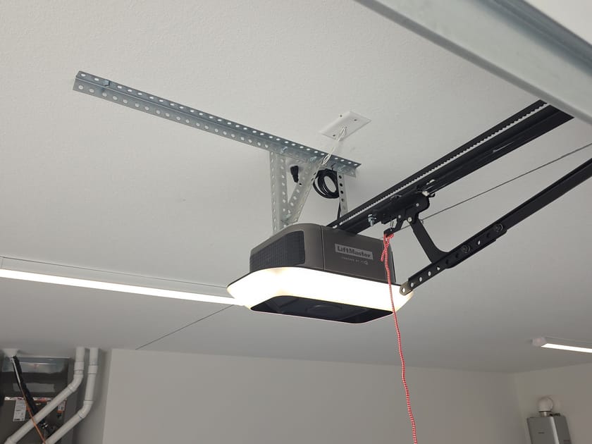 A garage door opener and its rail mounted to a garage ceiling, with the red emergency release cord hanging from the trolley.