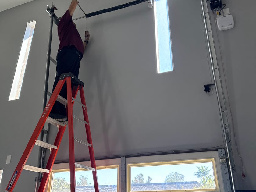 A technician on a red stepladder fitting the opener rail to the ceiling above a garage door.