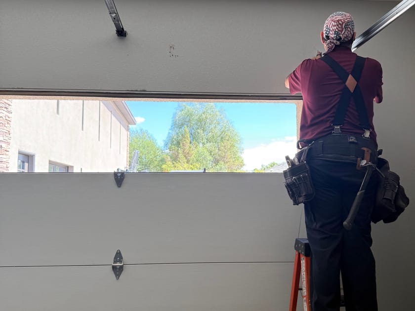 A Prestige Garage Door Services technician on a ladder, reaching the top panel of a garage door from inside the garage.