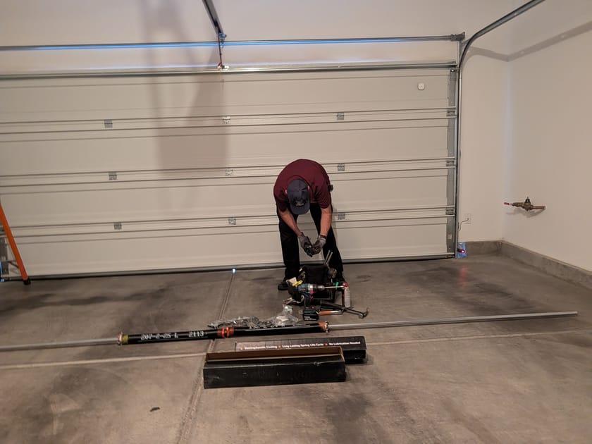 A technician kneeling on a garage floor assembling a garage door opener beside its rail sections and tools.