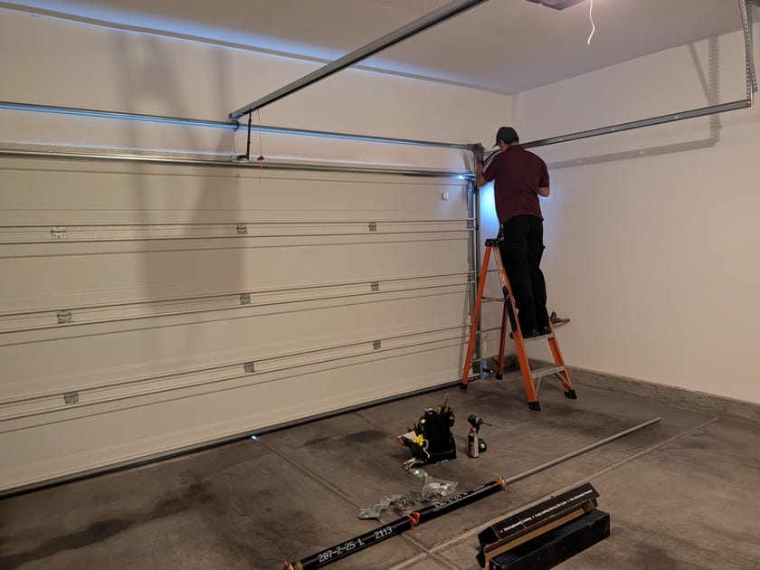 A technician on a stepladder fitting a panel of a new garage door, seen from inside the garage.