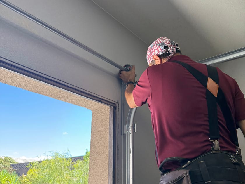 A technician working at the vertical track beside a part open garage door, with daylight in the opening.